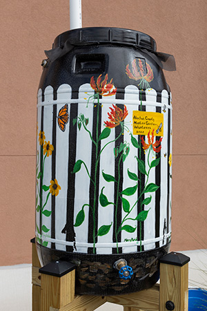 A large black barrel that has been converted into a rain barrel and painted with a white picket fence and flowers, a spigot at the bottom and its resting on a raised wooden stand