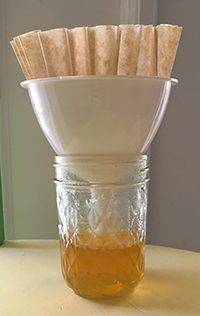 A white plastic funnel with a brown coffee filter nested inside, sitting on a glass jar that has yellow liquid in it