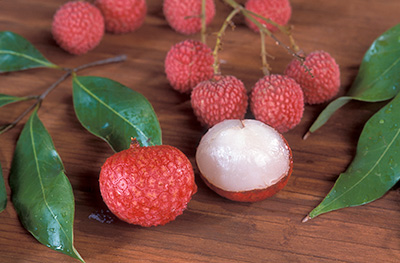 Red round and bumpy lychee fruits on a table with a few leaves. One fruit is peeled to expose the white flesh