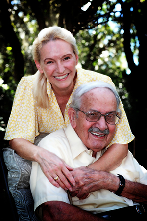 A woman with long blond hair standing behind and embracing a seated man with glasses and a mustache