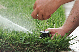 Two hands adjusting a irrigation sprinkler head as it sprays water onto grass and away from the sidewalk
