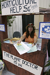 Woman sitting behind a booth with a sign reading Horticultural Help - Free! and The plant doctor is in