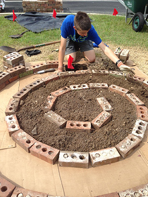 Young man kneeling over the beginning of a spiral garden: bricks laid in a spiral pattern on cardboard, with dirt filling in the spiral