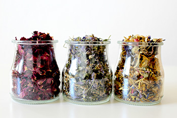 three small glass jars each with a different melange of dried herbs and flowers, the first being deep scarlet, the second being green and gray, and the third mainly brown with bits of purple