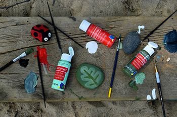 art supplies on a wooden table, plus two painted rocks one resembles a lady bug and the other is green with a leaf design