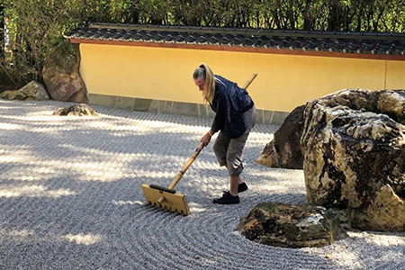 woman with long blond ponytail carefully raking a circular pattern into gravel around a large boulder