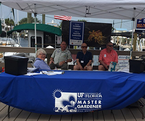 Three gentlemen and one woman sitting behind a table with a blue Master Gardener Volunteer logo, at a boat dock