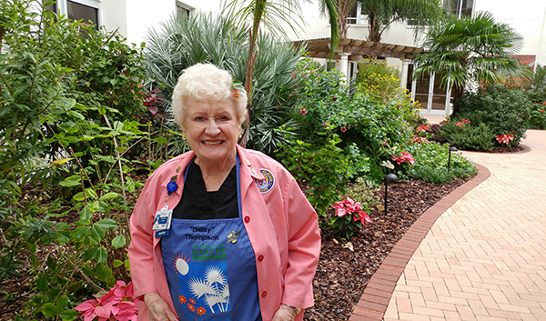 A smiling woman in a Master Gardener apron with the name Daisy stitched on it, standing in a garden with a brick patio