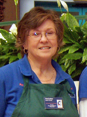 A bespectacled woman with short hair smiling shyling and wearing a green Master Gardener apron