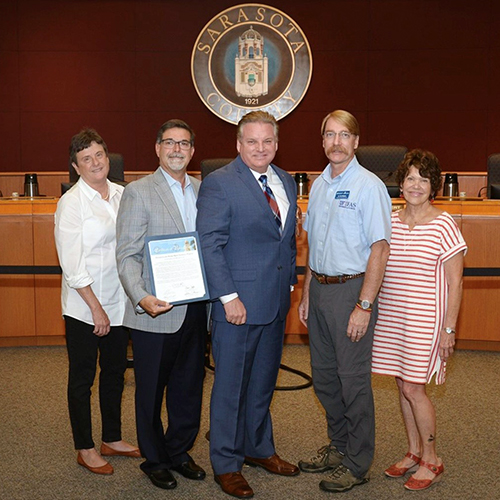 Three men, flanked by two women on each side, standing in front of the Sarasota County seal; one man is holding a certificate