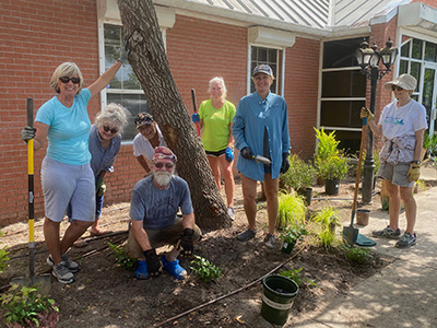 Seven volunteers pose with their garden tools as they work in a landscape in front of a brick building