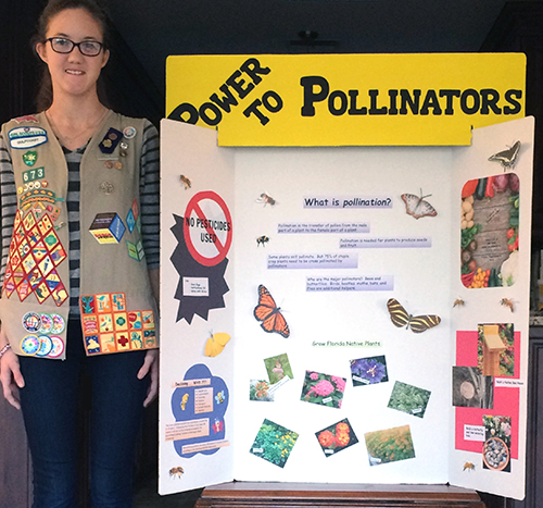 Girl with vest with many badges standing next to a poster display with the title Power to Pollinators