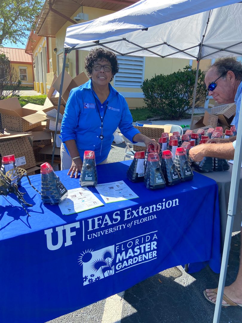 A smiling Master Gardener Volunteer standing behind outdoor table with mosquito traps to give away