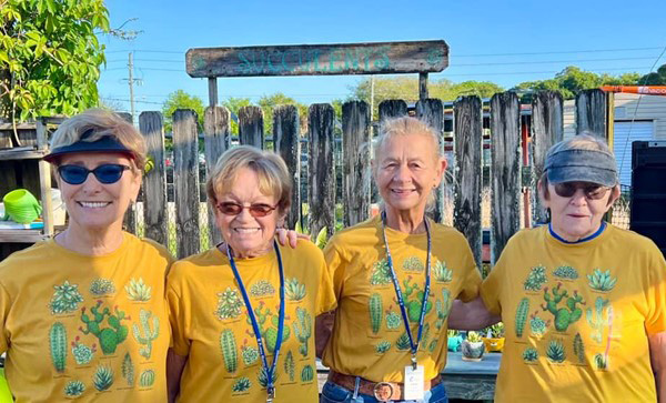 Four smiling women wearing matching yellow shirts, standing in a garden.