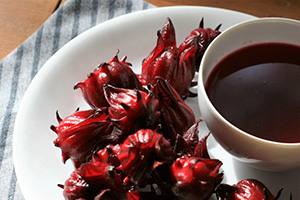 Several red waxy roselle hips sit next to a cup of dark red tea on a white plate.