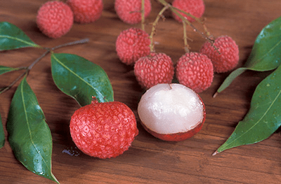 Red round and bumpy lychee fruits on a table with a few leaves. One fruit is peeled to expose the white flesh