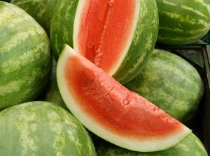A pile of green watermelons with one cut open to reveal the bright pinkish-red flesh inside.