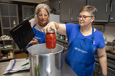 Two women stand over a huge pot on a stove. One is setting a glass jar of tomatoes in the pot.