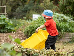 A small toddler attempts to pick up a huge yellow plastic bin in the garden. They have an adorable sun hat on.