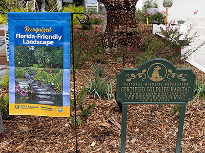 A mulched landscape bed displays a Florida-Friendly certification flag and a decorative metal sign